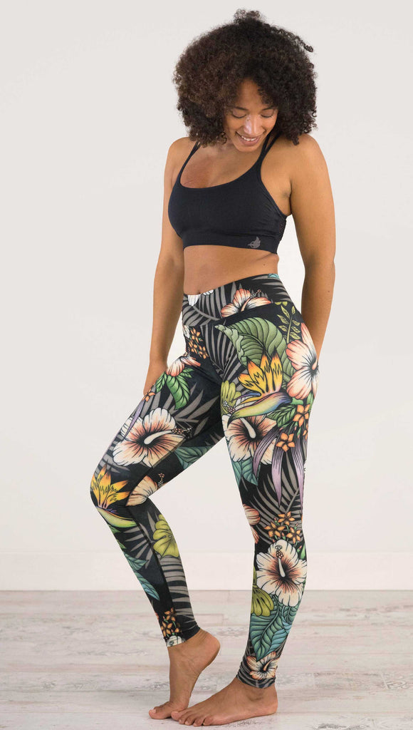 Full body side view of model wearing WERKSHOP Floral Night Full Length Triathlon Leggings. The artwork on the leggings has tropical flowers (bird of paradise, hibiscus and palm leaves) on a distressed black background.