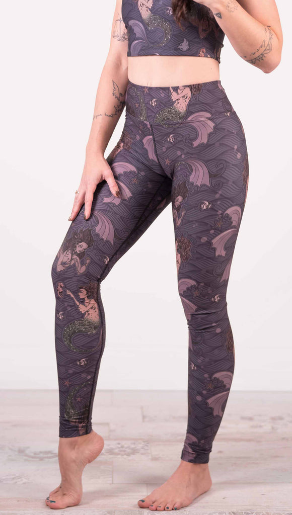 Model wearing WERKSHOP full length triathlon leggings with Original Mermaids artwork. The mermaids have small intricate details on the fins and are swimming with seahorses and angel fish over a dark blue background with waves.