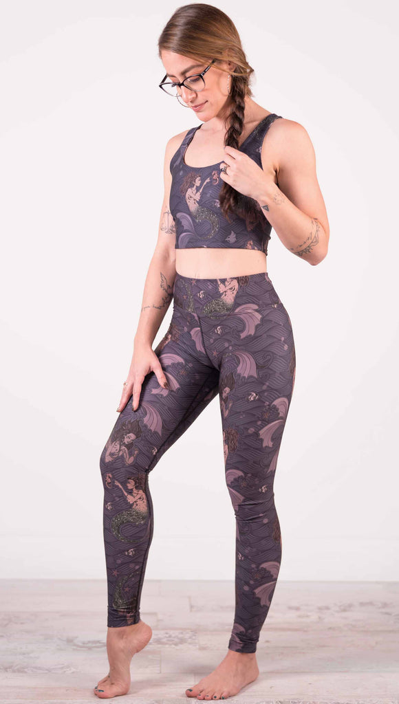 Model wearing WERKSHOP full length triathlon leggings with Original Mermaids artwork. The mermaids have small intricate details on the fins and are swimming with seahorses and angel fish over a dark blue background with waves.