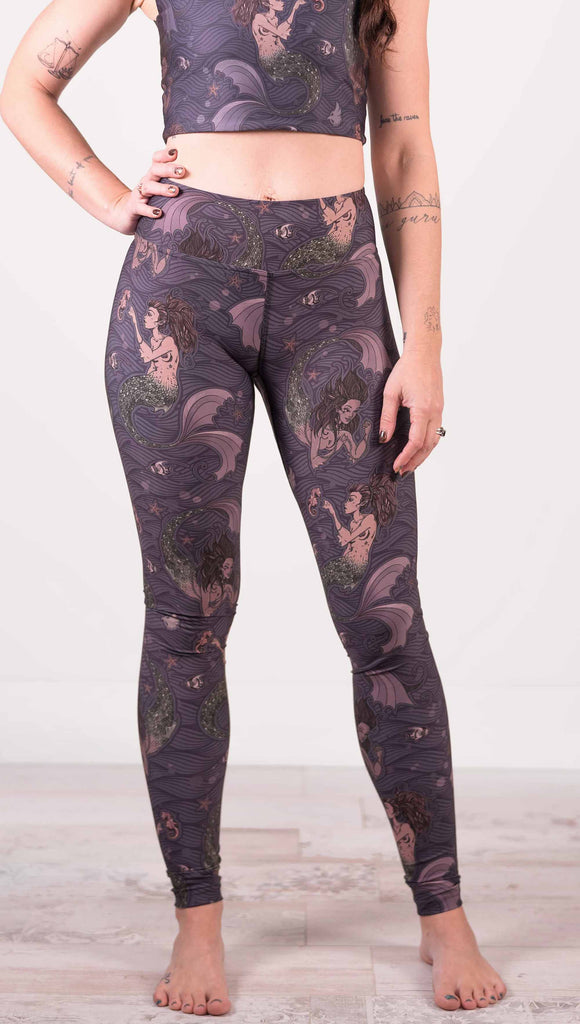 Model wearing WERKSHOP full length triathlon leggings with Original Mermaids artwork. The mermaids have small intricate details on the fins and are swimming with seahorses and angel fish over a dark blue background with waves.