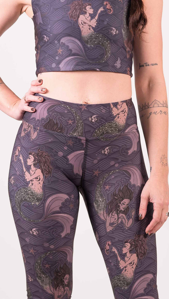 Model wearing WERKSHOP full length triathlon leggings with Original Mermaids artwork. The mermaids have small intricate details on the fins and are swimming with seahorses and angel fish over a dark blue background with waves.