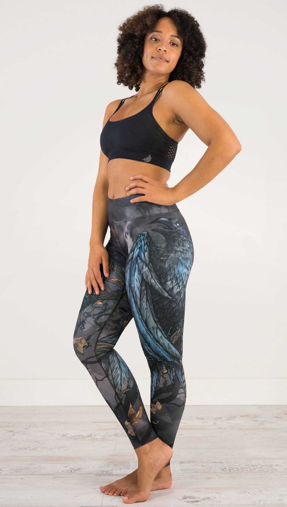 Full body side view of model wearing WERKSHOP Raven Full Length Triathlon Leggings. The artwork on the leggings is super dark shades of gray and blue with a large raven on the thigh and a dark and stormy moon in the background. Definitely spooky season vibes.