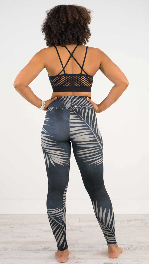 Full body back view of model wearing WERKSHOP Black Palms leggings. The leggings are printed with a mostly black watercolor effect with grayish palm frond silhouette wrapping the body.