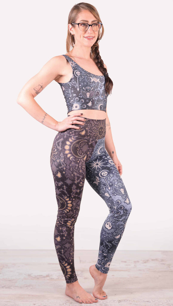 Model wearing WERKSHOP Zodiac and Tarot Mashed up Athleisure Leggings. The wearer’s right leg features zodiac themed artwork with the sun and moon with the moon phases, shooting stars and all 12 zodiac constellations in gold over a dark purple background. The left leg blue/tarot artwork has skulls, snakes, moons and the names of popular cards in the deck.