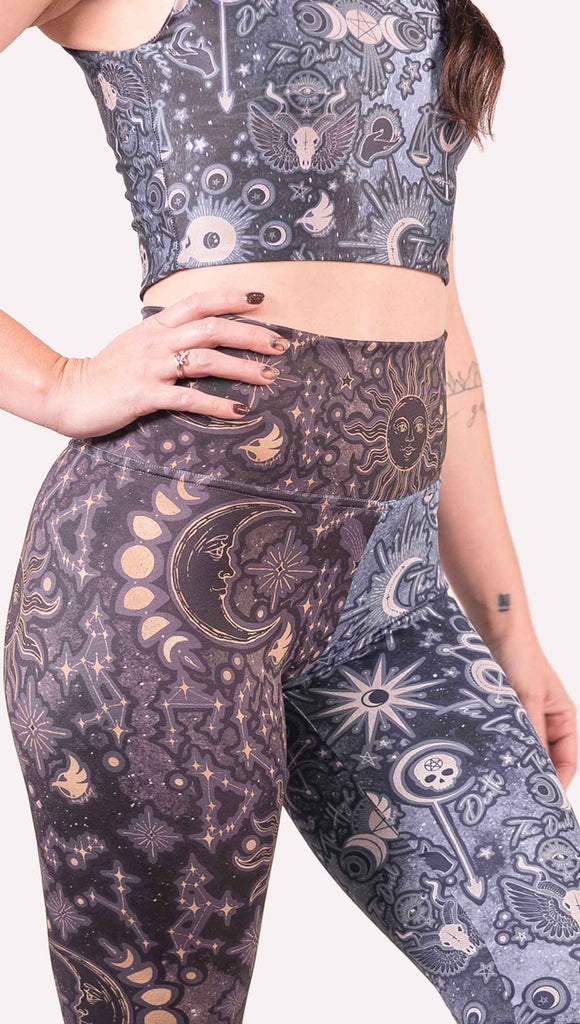 Model wearing WERKSHOP Zodiac and Tarot Mashed up Athleisure Leggings. The wearer’s right leg features zodiac themed artwork with the sun and moon with the moon phases, shooting stars and all 12 zodiac constellations in gold over a dark purple background. The left leg blue/tarot artwork has skulls, snakes, moons and the names of popular cards in the deck.