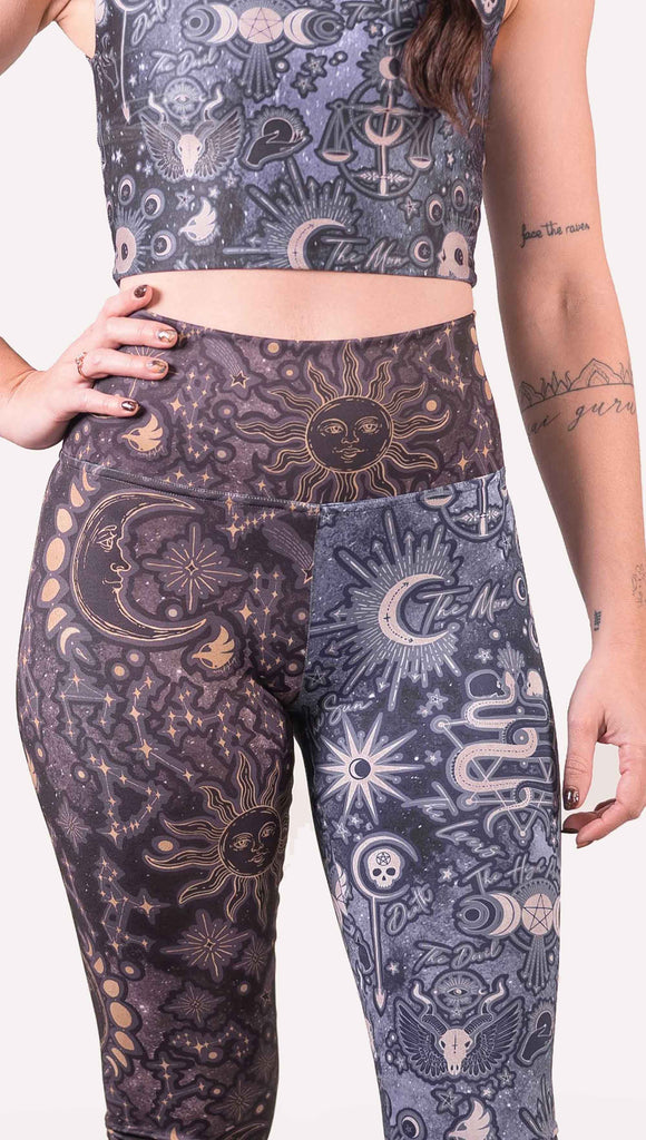 Model wearing WERKSHOP Zodiac and Tarot Mashed up Athleisure Leggings. The wearer’s right leg features zodiac themed artwork with the sun and moon with the moon phases, shooting stars and all 12 zodiac constellations in gold over a dark purple background. The left leg blue/tarot artwork has skulls, snakes, moons and the names of popular cards in the deck.