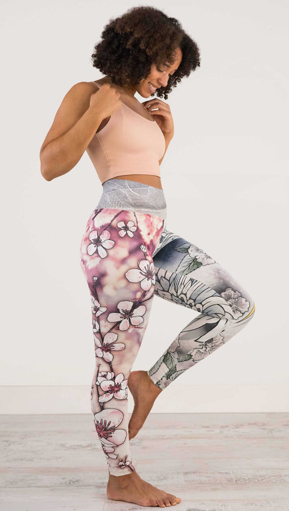 Full body side view of model wearing WERKSHOP Zen Mashup Athleisure Leggings. The leggigns are printed with pink cherry blossoms on the wearer's left leg and a graceful swooping crane on the opposite leg. There is also an abstract outline drawing of a koi fish on the waistband.