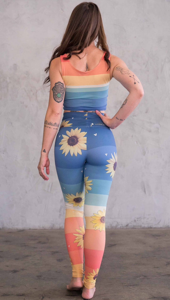 Full body back view of model wearing WERKSHOP Sunflower Athleisure leggings. The leggings have wide horizontal stripes with dark blue at the waistband, to aqua and pale green at the mid thigh leading to cream at the knee and orange and red tones to the ankle. There are large photo-real sunflowers and tiny hand sketched off-white hearts sprinkled throughout.