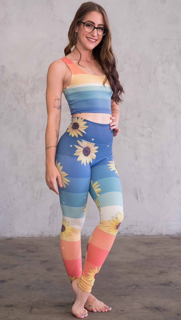 Full body front view of model wearing WERKSHOP Sunflower Athleisure leggings. The leggings have wide horizontal stripes with dark blue at the waistband, to aqua and pale green at the mid thigh leading to cream at the knee and orange and red tones to the ankle. There are large photo-real sunflowers and tiny hand sketched off-white hearts sprinkled throughout.