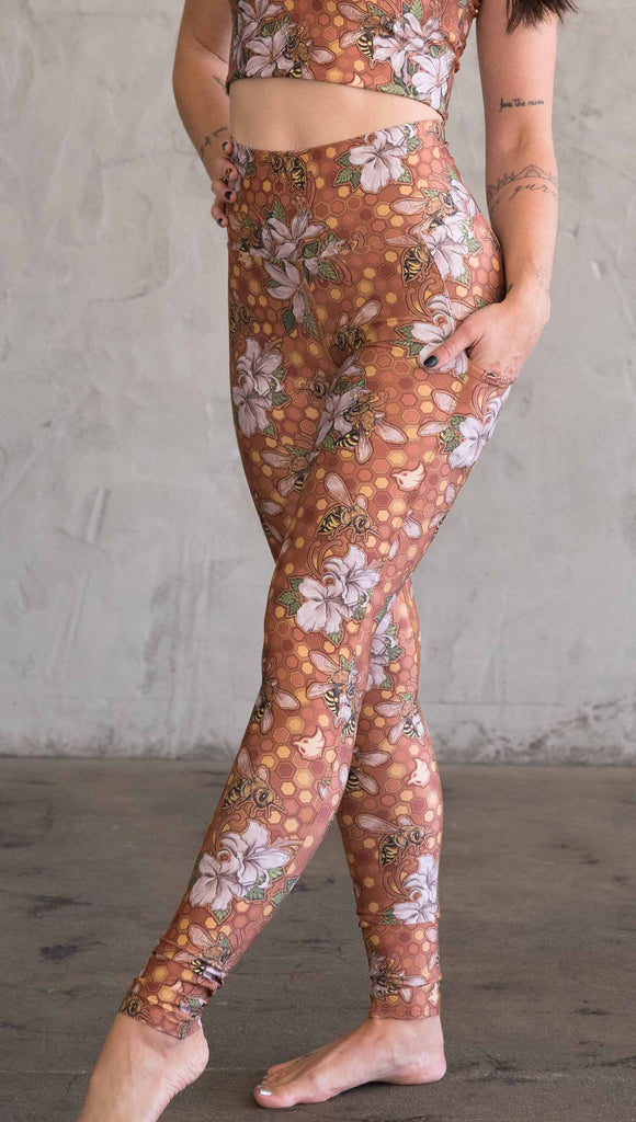 Side view of model wearing ultra lightweight "featherlight" leggings with clusters of honeybees and flowers. Her hand is in the side pocket that is big enough to hold a phone. 