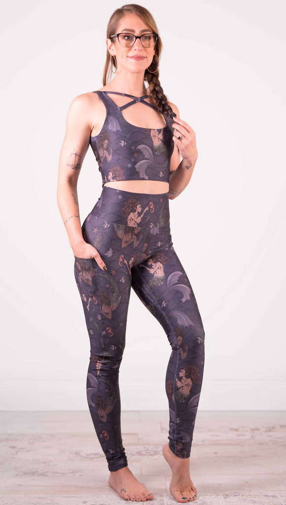 Full body front view of model wearing WERKSHOP featherlight leggings with Original Mermaids artwork. The mermaids have small intricate details on the fins and are swimming with seahorses and angel fish over a dark blue background with waves. The leggings have large pockets on each hip large enough to hold a phone.