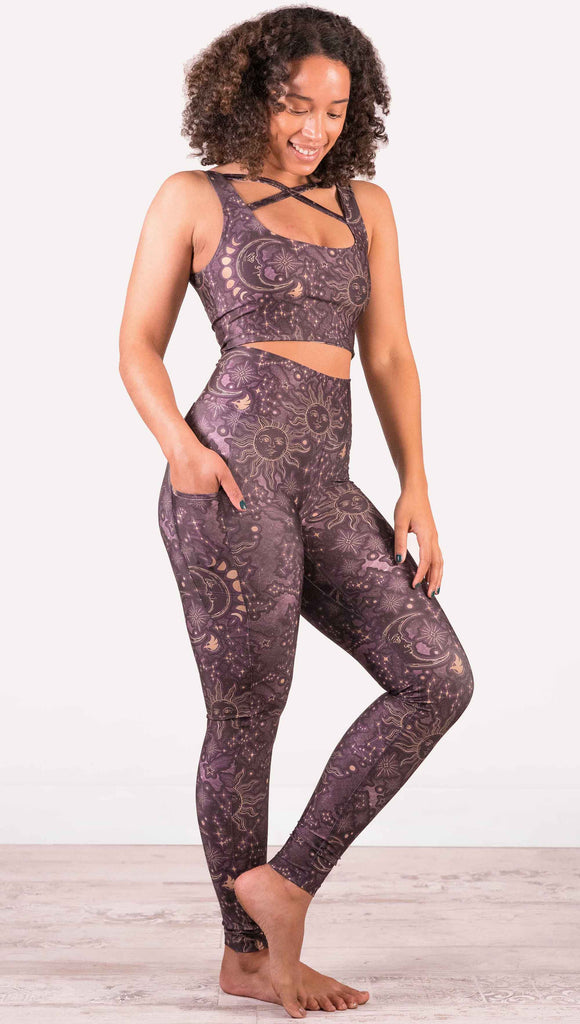 Model wearing WERKSHOP Zodiac Leggings. They are high waist and feature pockets on both legs. The zodiac themed artwork shows a hand-drawn sun and moon with the moon phases, shooting stars and all 12 zodiac constellations in gold over a dark purple background.