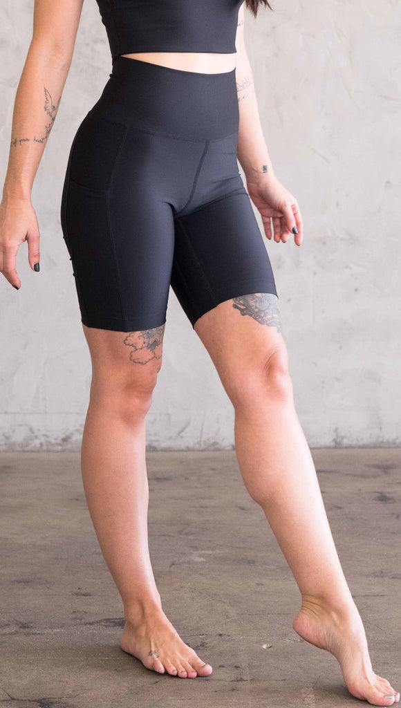 Waist down view of model wearing WERKSHOP bicycle length shorts in featherlight fabric. Black color with side cell phone pockets and a small eagle logo on the wearers left side