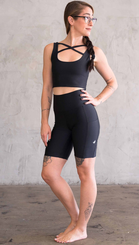 Full body front view of model wearing WERKSHOP bicycle length shorts in featherlight fabric. Black color with side cell phone pockets and a small eagle logo on the wearers left side