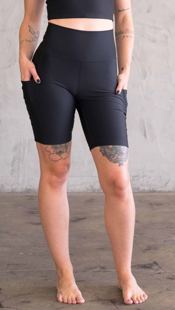 Waist down view of model wearing WERKSHOP bicycle length shorts in featherlight fabric. Black color with side cell phone pockets and a small eagle logo on the wearers left side