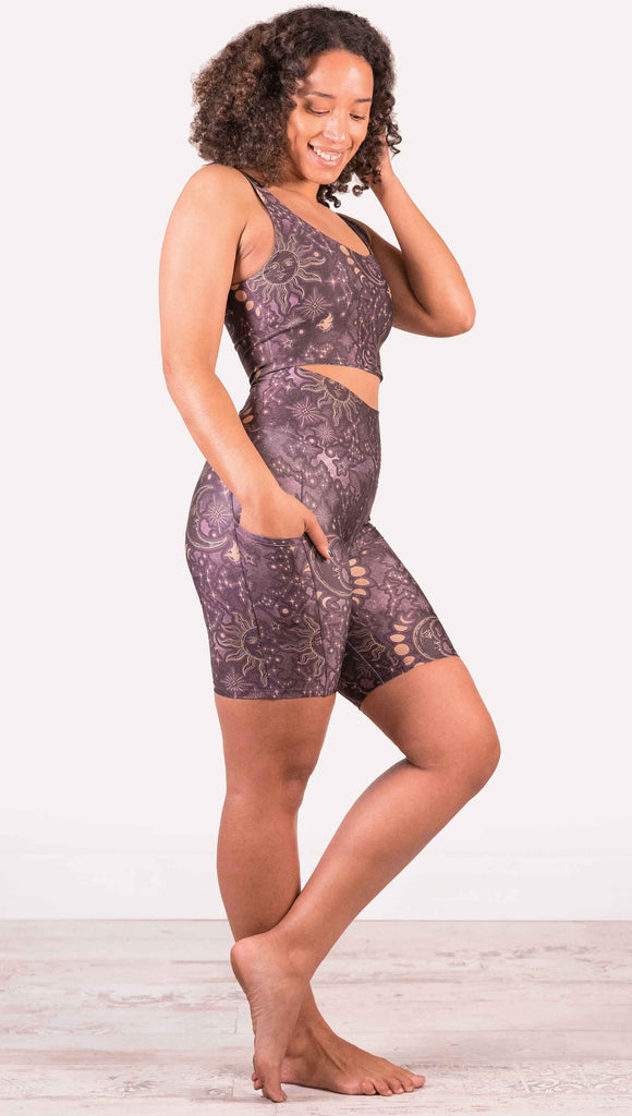 Model wearing WERKSHOP Zodiac Bicycle Length Shorts. They are high waist and feature pockets on both legs. The zodiac themed artwork shows a hand-drawn sun and moon with the moon phases, shooting stars and all 12 zodiac constellations in gold over a dark purple background. The length of the shorts hit in the mid thigh, above the knee.