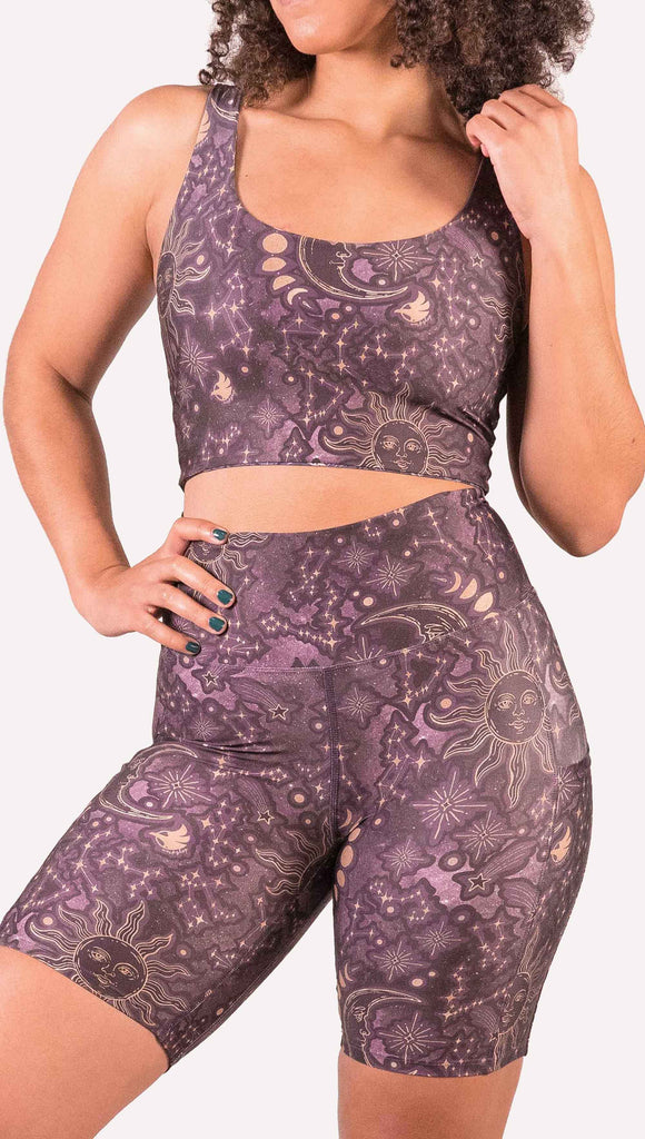 Model wearing WERKSHOP Zodiac Bicycle Length Shorts. They are high waist and feature pockets on both legs. The zodiac themed artwork shows a hand-drawn sun and moon with the moon phases, shooting stars and all 12 zodiac constellations in gold over a dark purple background. The length of the shorts hit in the mid thigh, above the knee.