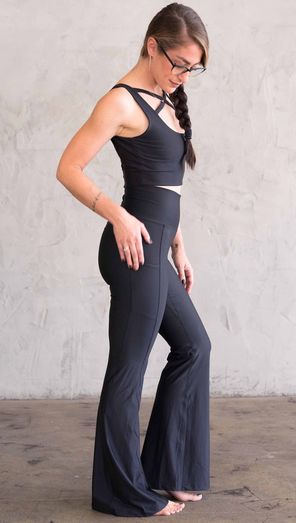 Full body side view of model wearing WERKSHOP Featherlight Bell Bottoms in solid black. The bells have a small reflective eagle logo on the wearer's left side pocket.