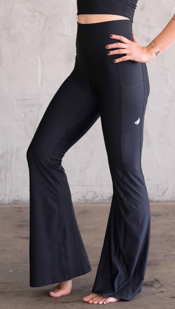 Waist down view of model wearing WERKSHOP Featherlight Bell Bottoms in solid black. The bells have a small reflective eagle logo on the wearer's left side pocket.