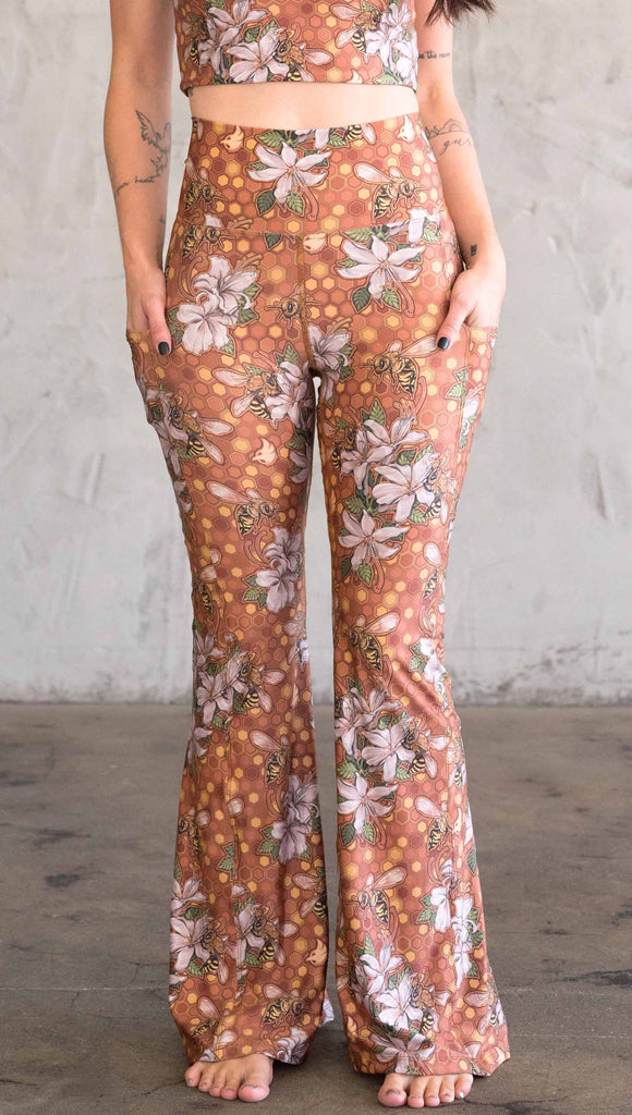 Front view of model wearing WERKSHOP Honeybee Bell Bottoms. The leggings have clusters of honeybees and flowers and phone pockets on each leg