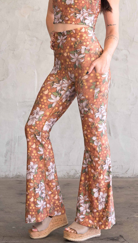 Side view of model wearing WERKSHOP Honeybee Bell Bottoms. The leggings have clusters of honeybees and flowers and phone pockets on each leg