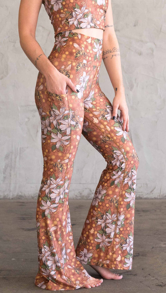 Side view of model wearing WERKSHOP Honeybee Bell Bottoms. The leggings have clusters of honeybees and flowers and phone pockets on each leg