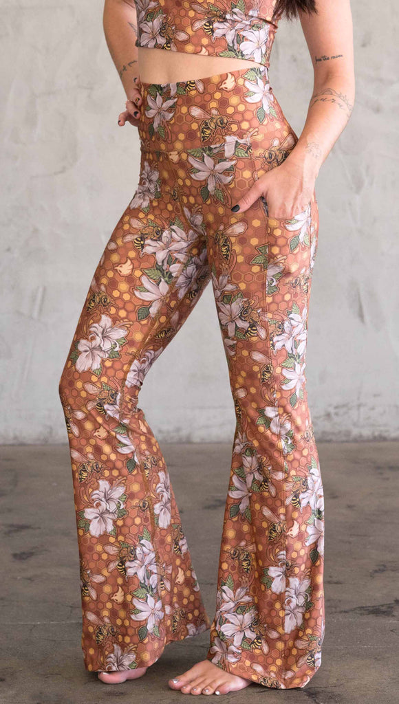 Side view of model wearing WERKSHOP Honeybee Bell Bottoms. The leggings have clusters of honeybees and flowers and phone pockets on each leg