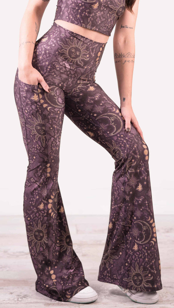 Model wearing WERKSHOP Zodiac Bell Bottoms. They are high waist and feature pockets on both legs. The zodiac themed artwork shows a hand-drawn sun and moon with the moon phases, shooting stars and all 12 zodiac constellations in gold over a dark purple background. Our model is 5’2” and this images shows her wearing the bells with sneakers. The hem of the pants are hitting the floor.