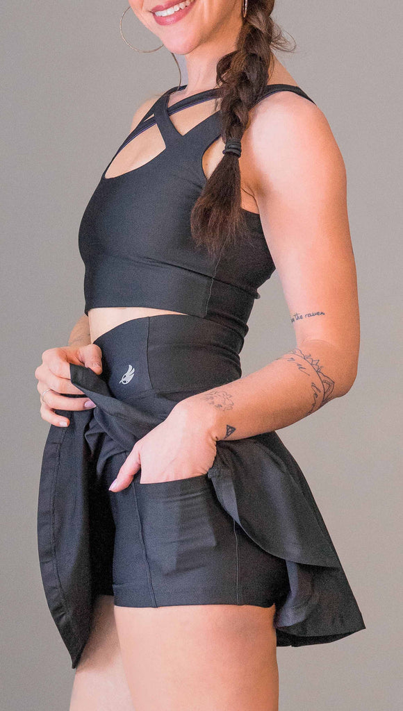 Zoomed in view of model wearing WERKSHOP Tennis Skirt, with the skirt lifted up to show the shorts and pockets underneath. In solid black color