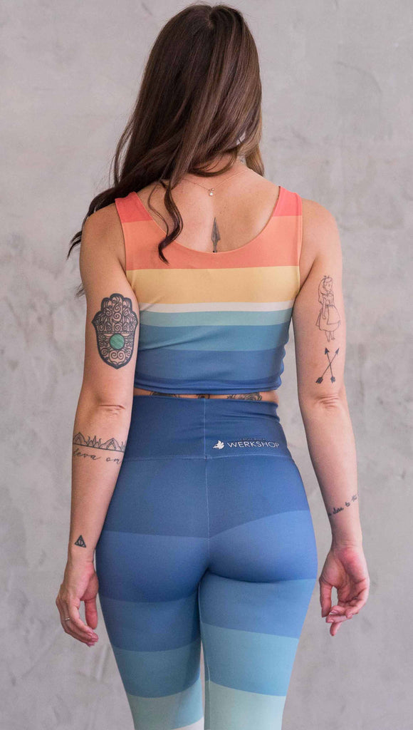 Back view of model wearing WERKSHOP Sunflower Rainbow Top. The top has vintage colored rainbow stripes on both sides. The stripes start orange at the shoulders, yellow and cream across the chest and then blue at the sweep. One side also has sunflowers and small cream colored hearts.