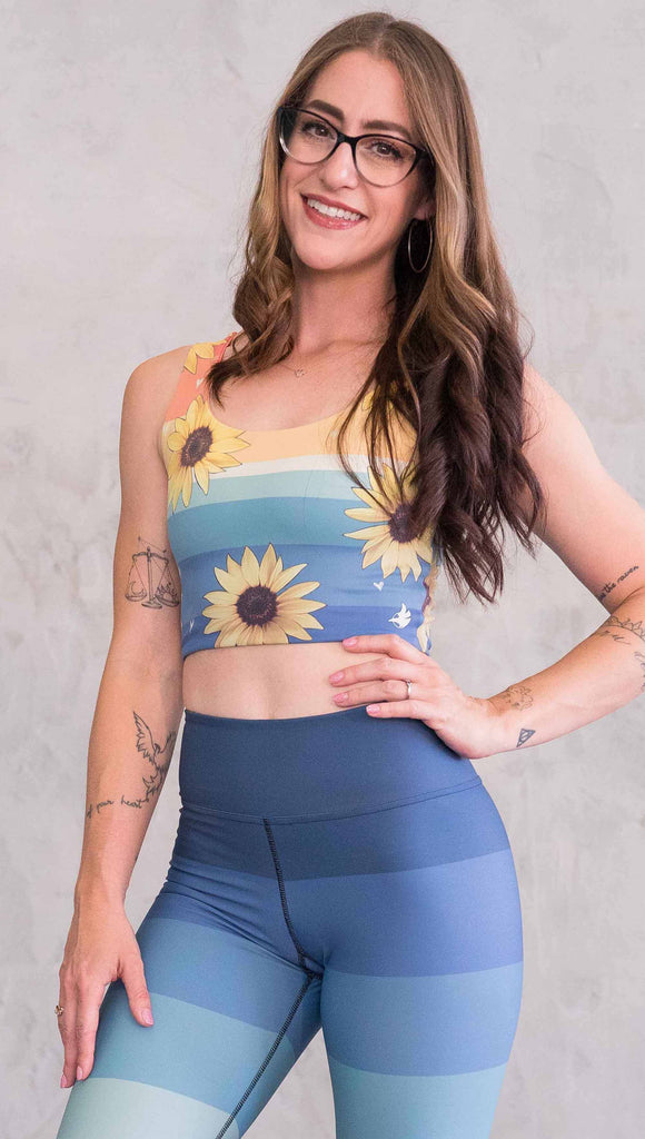 Front view of model wearing WERKSHOP Sunflower Rainbow Top. The top has vintage colored rainbow stripes on both sides. The stripes start orange at the shoulders, yellow and cream across the chest and then blue at the sweep. One side also has sunflowers and small cream colored hearts.