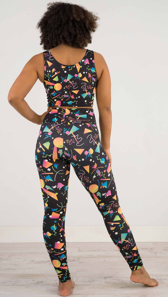 Full body view of model wearing WERKSHOP white confetti athleisure leggings. The artwork on the leggings shows multi-colored circles, scribbles and triangles over a black background.