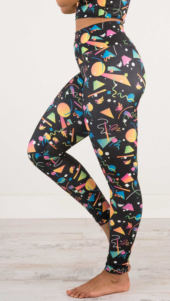 Waist down view of model wearing WERKSHOP white confetti athleisure leggings. The artwork on the leggings shows multi-colored circles, scribbles and triangles over a black background.