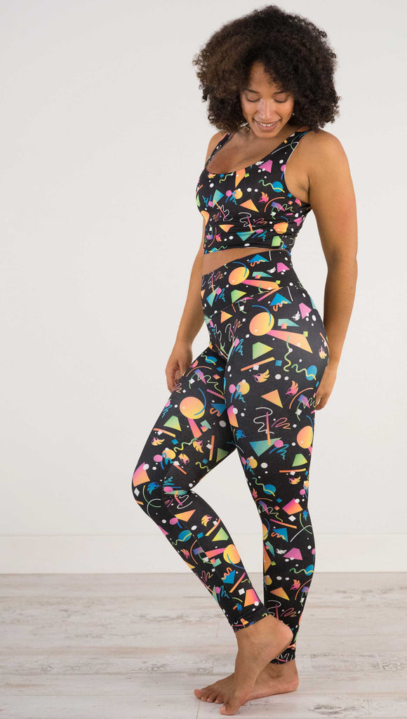 Full body view of model wearing WERKSHOP white confetti athleisure leggings. The artwork on the leggings shows multi-colored circles, scribbles and triangles over a black background.