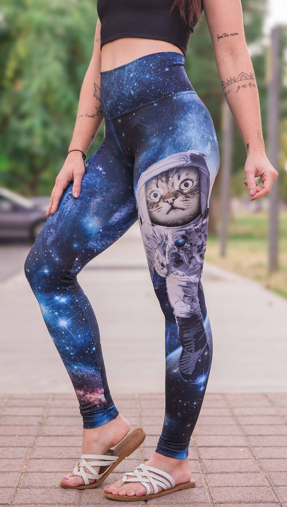 Model wearing Catstronaut Leggings. The leggings are printed with a cat in an astronaut suit on the wearer's left leg. The galaxy background is a deep cobalt blue with nebula swirls and shooting stars.