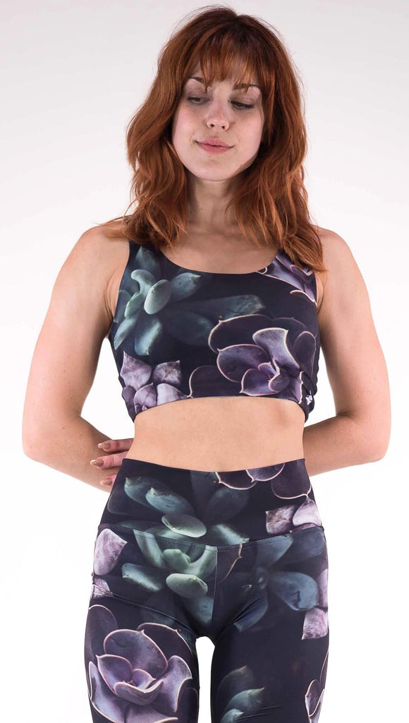 Front view of model wearing the Midnight Garden reversible top. This is in the Midnight Garden side, it is a black crop top with green and purple succulent plants throughout