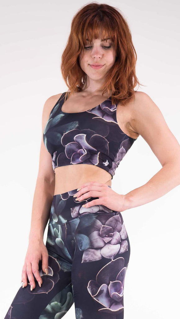 Left view of model wearing the Midnight Garden reversible top. This is in the Midnight Garden side, it is a black crop top with green and purple succulent plants throughout