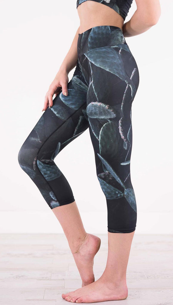 Enhanced left view of model wearing black capri leggings with dark green cacti plants throughout