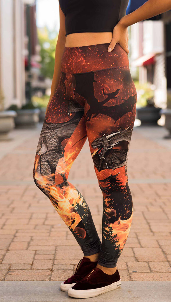 Model wearing WERKSHOP Spitfire Leggings. The artwork on the leggings features a detailed fire breathing dragon blowing flames into a forest with a bright red sky filled with floating embers and silhouettes of more dragons flying in the background.