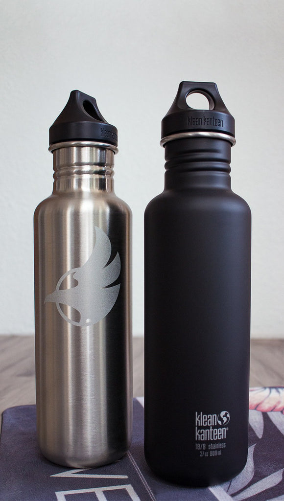 Black water bottle with Eagle Rock Werkshop logo next to silver water bottle with Eagle Rock Werkshop logo resting on a purple yoga mat with Eagle Rock Werkshop logo.