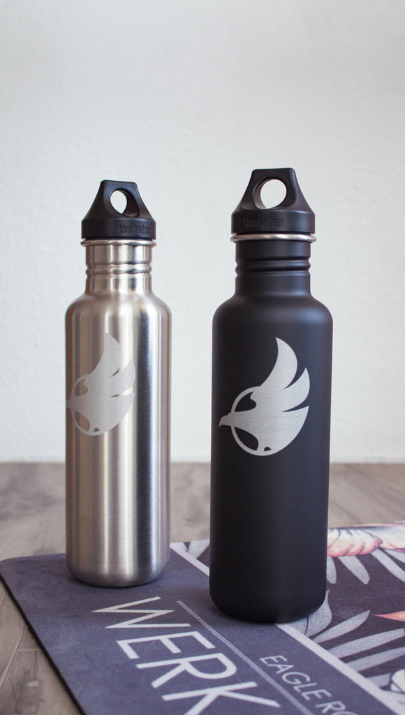 Black water bottle with Eagle Rock Werkshop logo next to silver water bottle with Eagle Rock Werkshop logo resting on a purple yoga mat with Eagle Rock Werkshop logo.