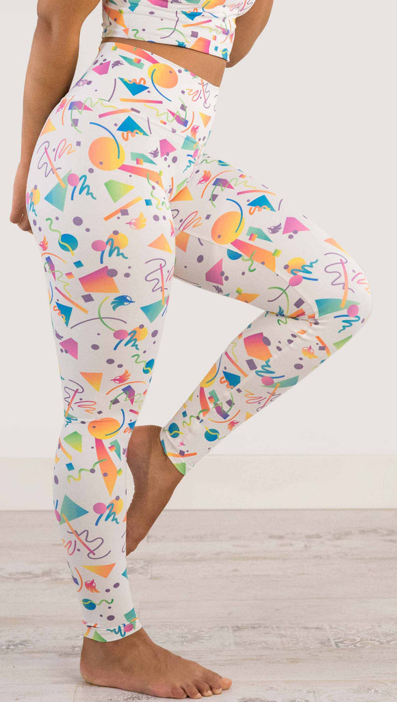 Waist down view of model wearing WERKSHOP white confetti athleisure leggings. The artwork on the leggings shows multi-colored circles, scribbles and triangles over a white background.