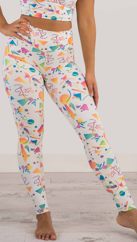 Waist down view of model wearing WERKSHOP white confetti athleisure leggings. The artwork on the leggings shows multi-colored circles, scribbles and triangles over a white background.